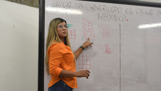 Se ve a la profesora Geraldin Giraldo frente a un tablero, señalando un esquema dibujado en marcador rojo mientras lo explica a sus estudiantes.