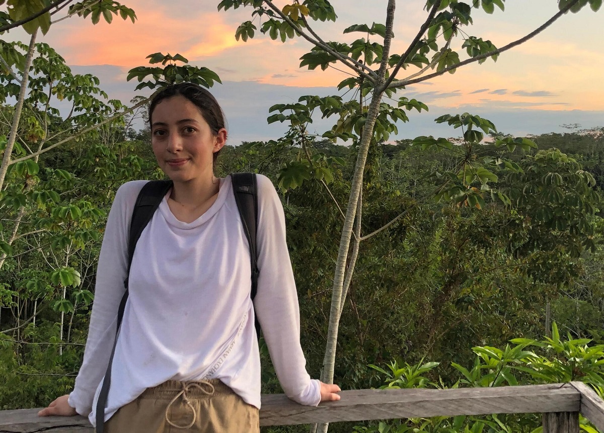 María Alejandra Jiménez, se encuentra en un bosque tropical. Viste una camisa blanca de manga larga, pantalones marrones y una mochila negra y el cielo está despejado y azul, con algunas nubes tenues