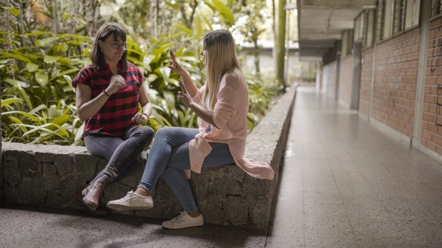 Dos mujeres se comunican en Lengua de Señas Colombiana, sentadas en el muro de una jardinera en un ambiente universitario. 