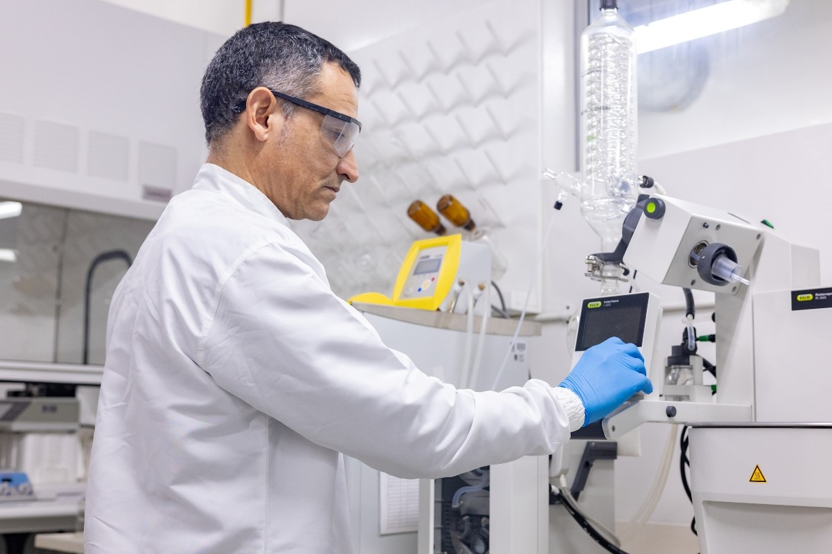 La imagen muestra al Jahir Orozco Holguín con gafas de seguridad, vistiendo una bata blanca, trabajando en un laboratorio. Está de pie frente a un equipo y tiene un guante azul en su mano derecha con la que está tocando la pantalla del equipo