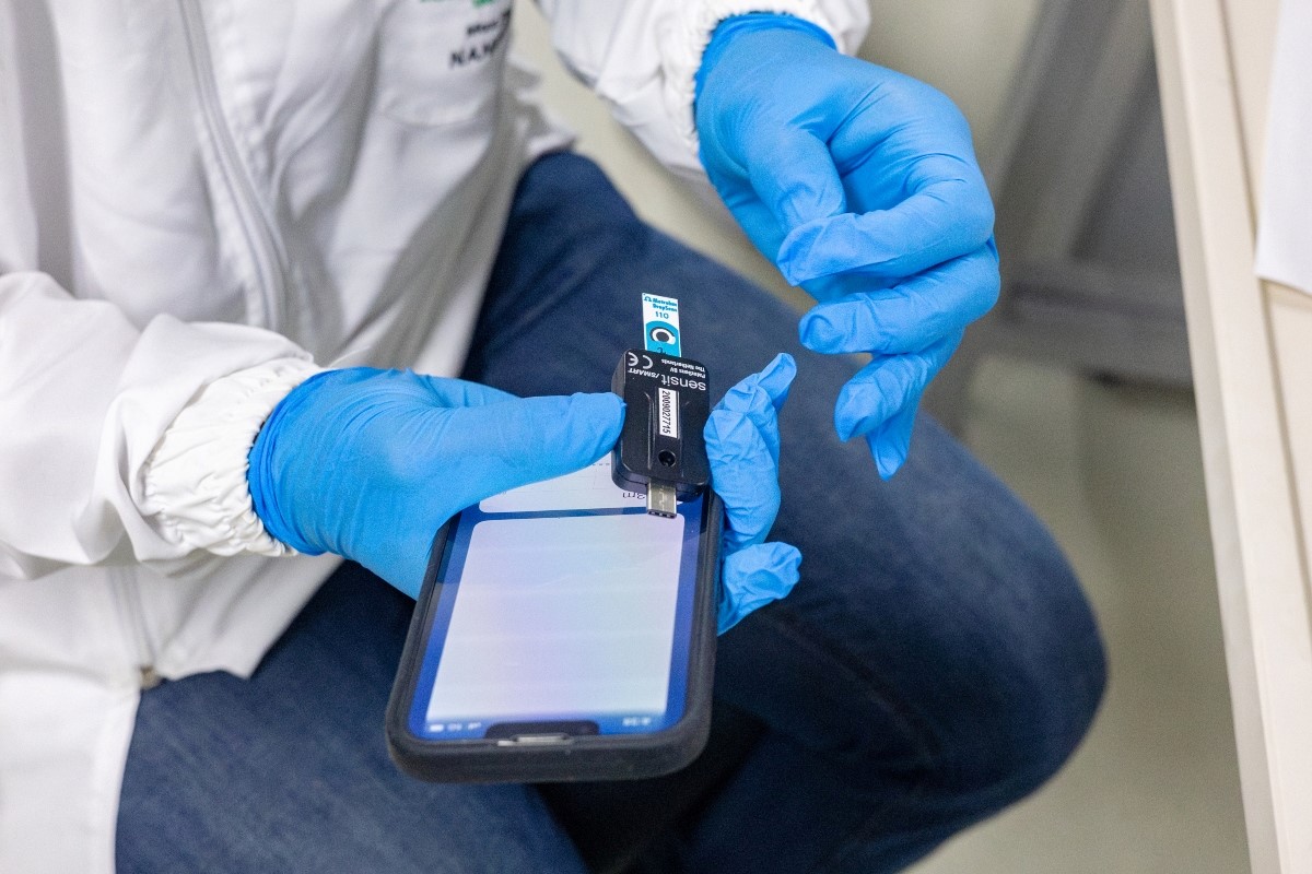 la imagen muestra una persona vestida con una bata blanca, jeans azules oscuros y guantes azules, la persona está sentada en una silla y sostiene en sus manos un dispositivo de Nanobioingeniería y un teléfono inteligente