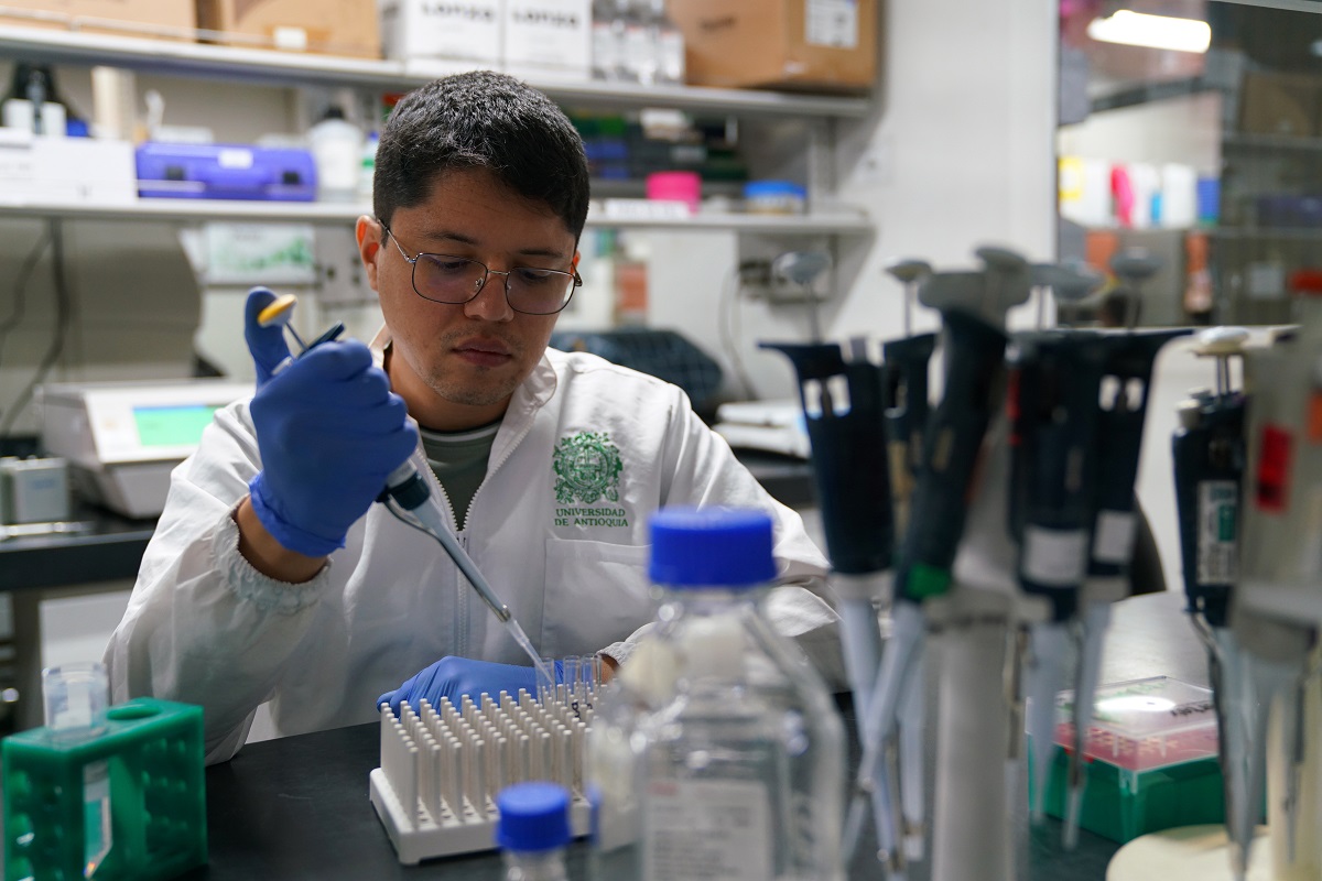 La foto muestra a un científico UdeA trabajando en un laboratorio. Está usando un equipo para dispensar líquido en tubos de ensayo. Lleva guantes de latex azules y una bata blanca con el logo de la UdeA en el lado izquierdod el pecho.