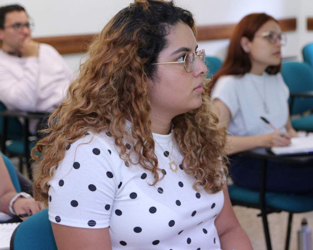 Primer plano de una estudiante en el salón de clases. Mira hacia la derecha, donde está el docente y el tablero.