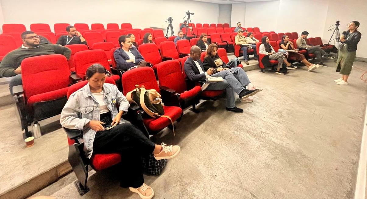 La imagen muestra a personas sentadas en un auditorio con sillas rojas. Varias están tomando notas o usando celulares. Al fondo hay cámaras sobre trípodes y hacia la derecha se encuentra una mujer de pie moderando el taller.