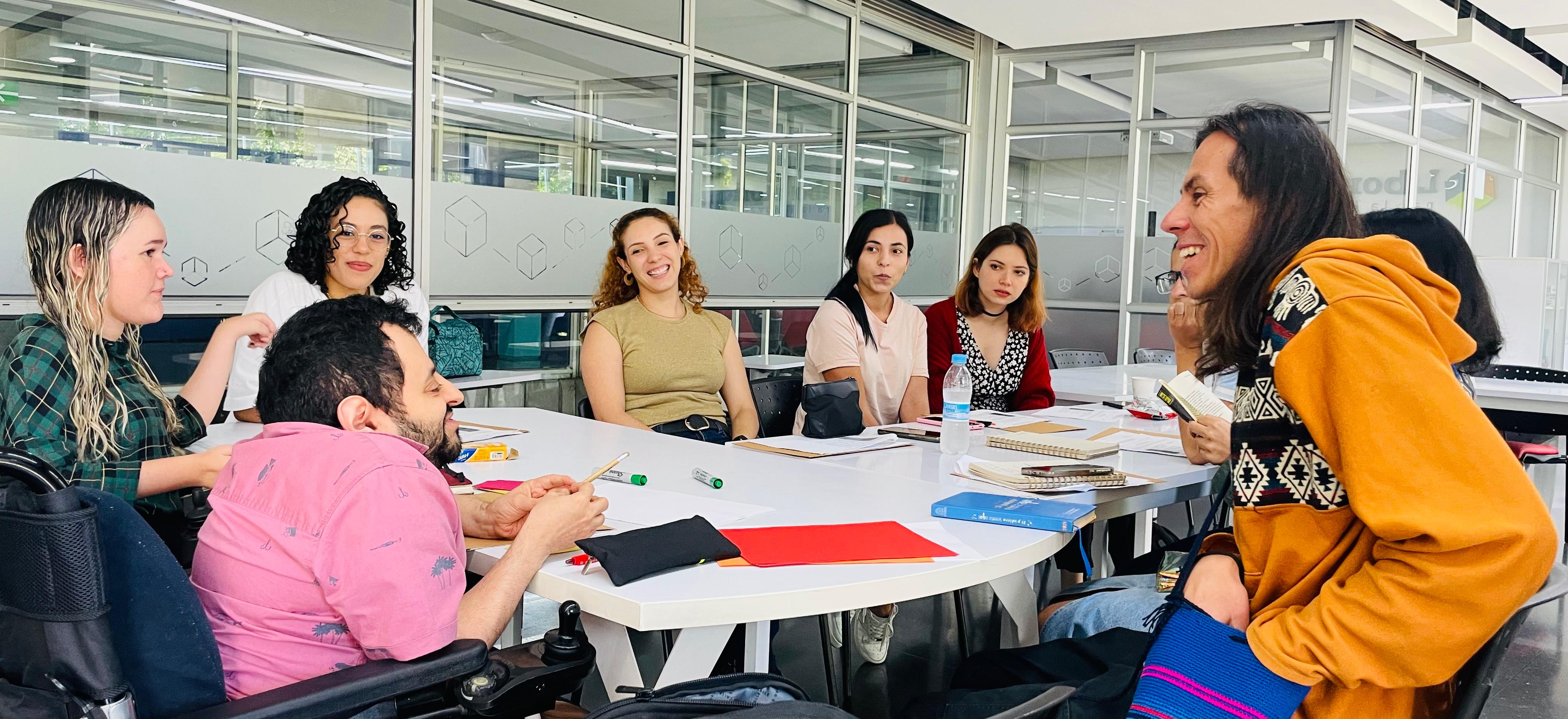 En la imagen se observa a un grupo de personas reunidas alrededor de una mesa, participando activamente en una conversación dentro de una dinámica de trabajo colaborativo.
