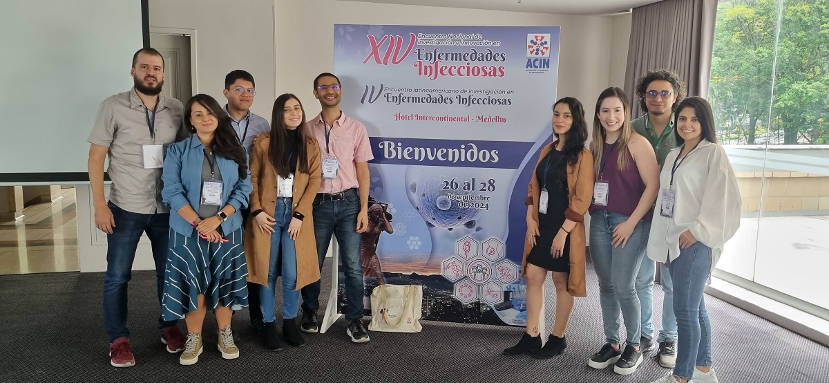 Esta foto muestra a un grupo de personas en un evento llamado "Encuentro Nacional de Investigación e Innovación en Enfermedades Infecciosas". Están de pie frente a un cartel que anuncia el evento. La gente está sonriendo y disfrutando del evento