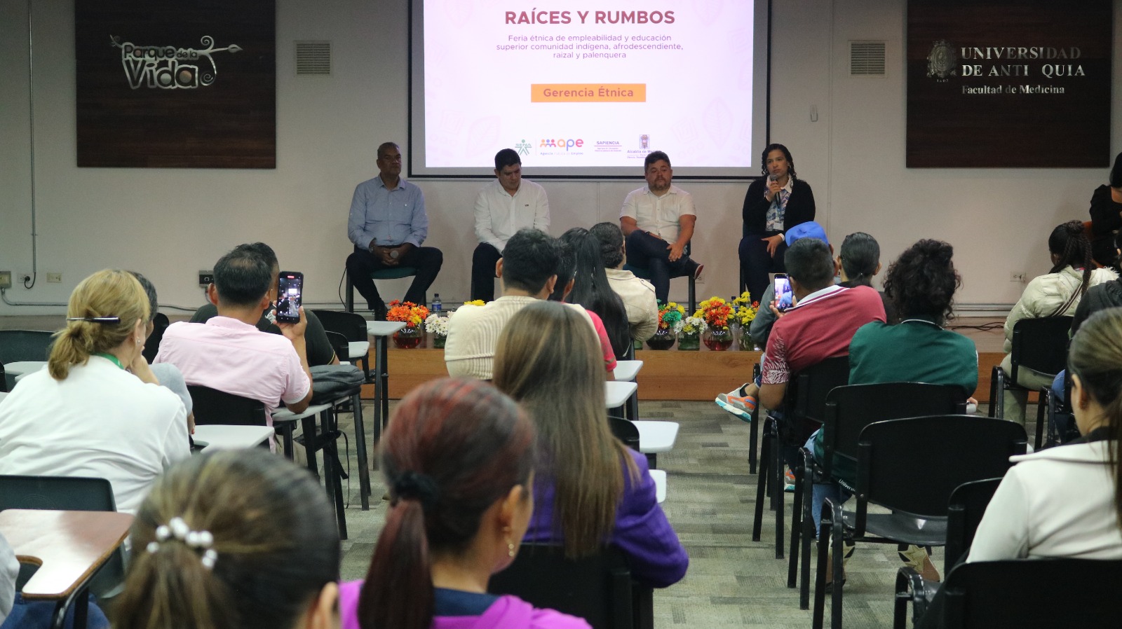 Foto: Cuatro personas aparecen sentadas en la tarima, frente a un grupo de personas sentadas en sillas en un auditorio.