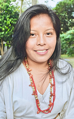 Profesora indígena wayuunaiki de largo cabello, con vestido tradicional blanco y collares rojos, sonriente.